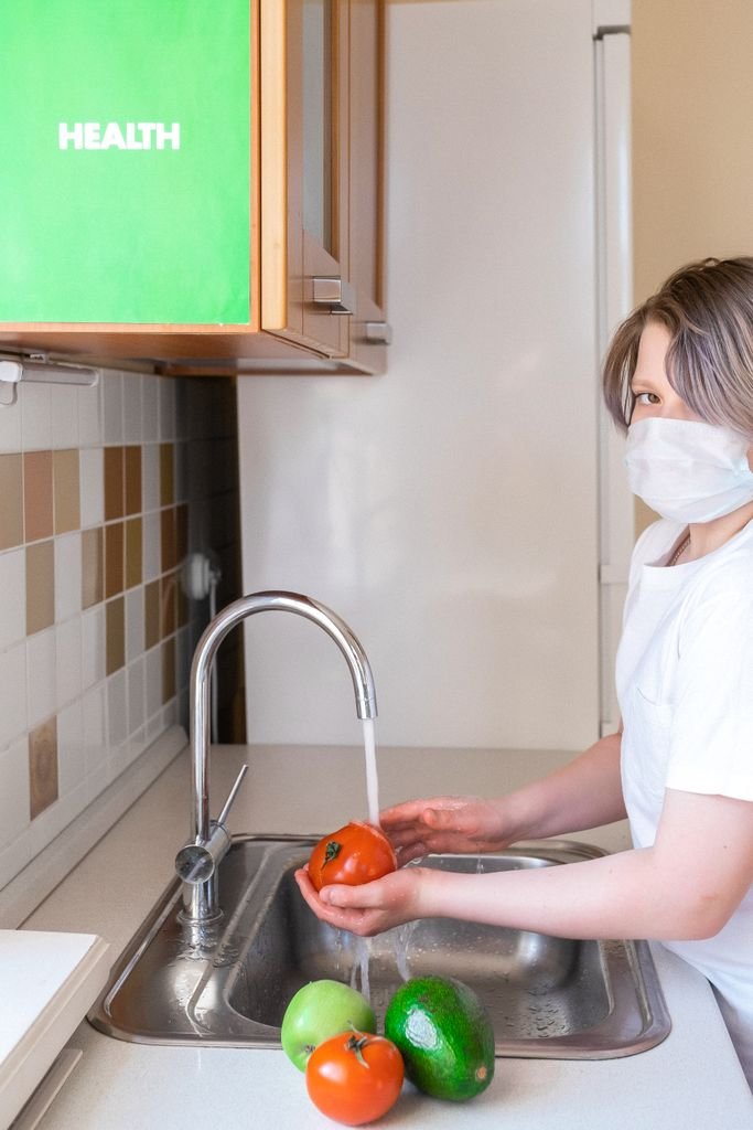 An adult wearing a mask washes vegetables in a kitchen sink for health safety.