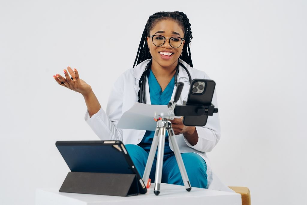 Black female doctor in virtual consultation, smiling at screen with stethoscope, tablet, and phone.